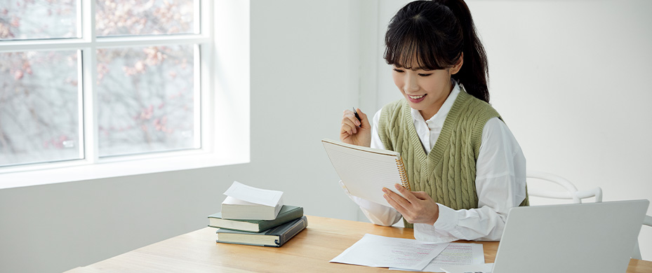 창가 책상에 앉은 여성이 미소를 지으며 손에 든 노트패드에 펜으로 글을 적고 있는 모습